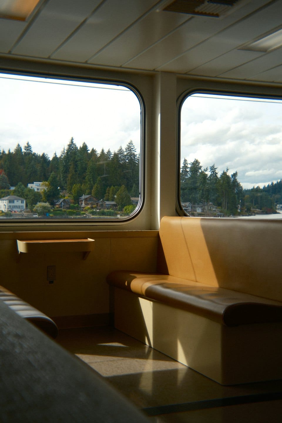 Two large ferry windows overlook a forested shoreline with houses.
