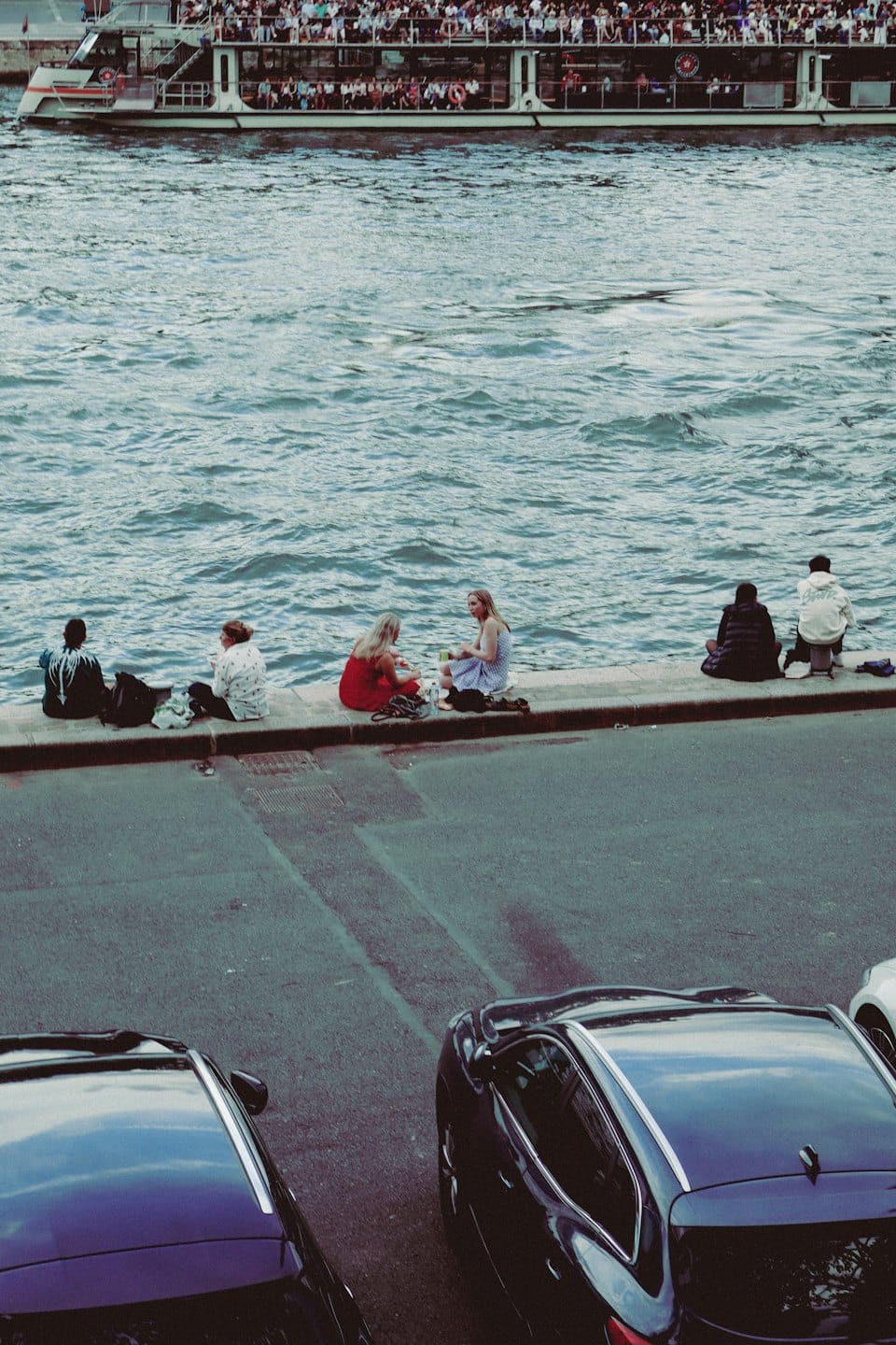 People sit by a river while a tourist boat passes.